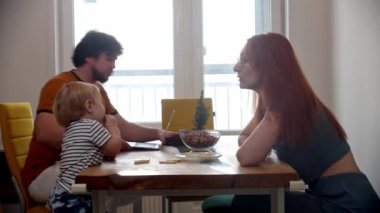 Masanın başında oturan küçük çocuklu bir aile. Babası dizüstü bilgisayarla çalışıyor ve annesi de bebeğine bakıyor.