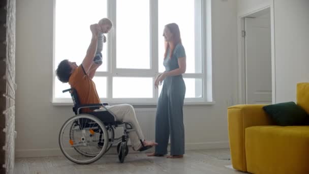 Hombre en silla de ruedas jugando con su pequeño bebé cerca del alféizar de la ventana y su esposa mirándolos — Vídeos de Stock