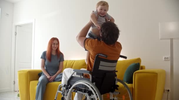 Mężczyzna na wózku inwalidzkim bawiący się swoim dzieckiem i uśmiechniętą żoną oglądającą je — Wideo stockowe
