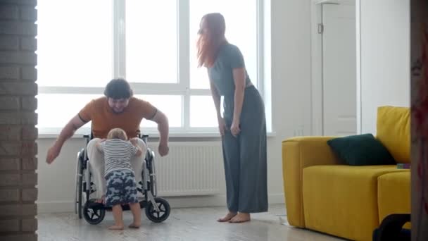 Küçük bir bebek ve annesi tekerlekli sandalyedeki adama koşuyorlar. Kadın kocasını öpüyor ve gülümsüyor. — Stok video