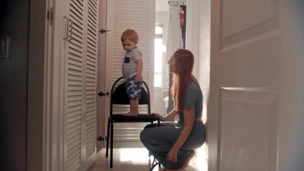 Ein kleines Baby steht auf dem Stuhl und wirft einen Kleiderbügel aus dem Kleiderschrank und seine Mutter legt ihn zurück — Stockvideo