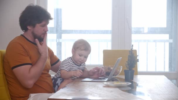 Solo padre trabaja en su portátil y su pequeño bebé se sienta en sus rodillas — Vídeos de Stock