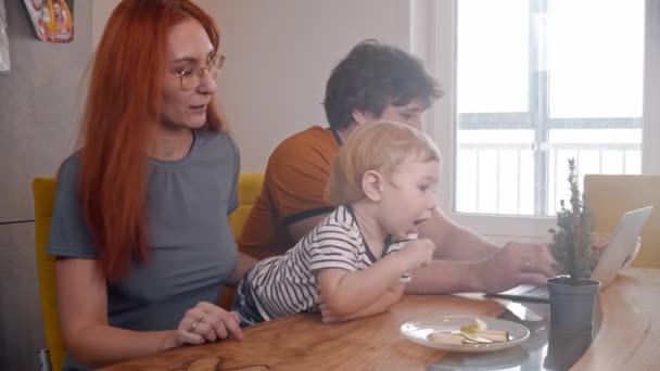 Hombre estresante tratando de trabajar con su portátil mientras su bebé se arrastra sobre la mesa - su esposa se lleva al bebé y lo besa en la mejilla — Vídeos de Stock