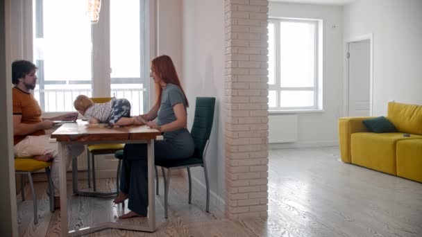 Hombre freelancer trabaja usando su portátil y su esposa y el bebé sentado en el otro lado de la mesa y luego alejarse en otra habitación — Vídeos de Stock