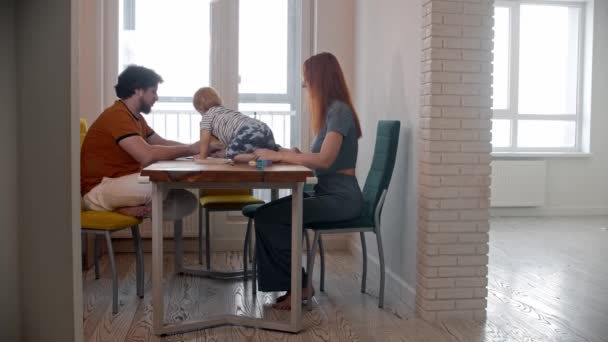 Gestresster Mann versucht mit seinem Laptop zu arbeiten, während sein Baby auf dem Tisch krabbelt - seine Frau nimmt das Baby mit und geht in ein anderes Zimmer — Stockvideo
