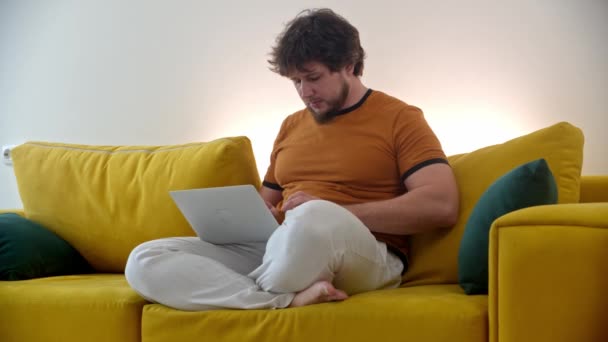 Homme pigiste assis sur un canapé jaune travaillant à la maison à l'aide d'un ordinateur portable — Video