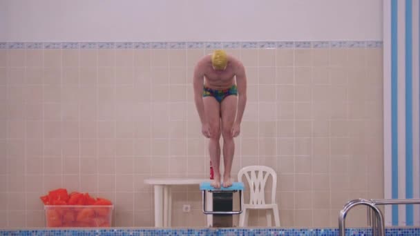 Ein Mann mit gelber Mütze springt im Schwimmbad von der Tribüne — Stockvideo