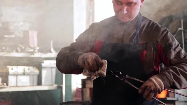 The blacksmith holds the cooled metal detail with tongs and wipes it with a rag — ストック動画