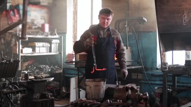 男の鍛冶屋は炉から赤い加熱された金属の詳細を取り出し、水のバケツで冷却します — ストック動画