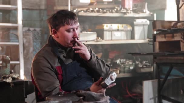 一个人坐在铁匠车间里抽烟和拿着锤子 — 图库视频影像