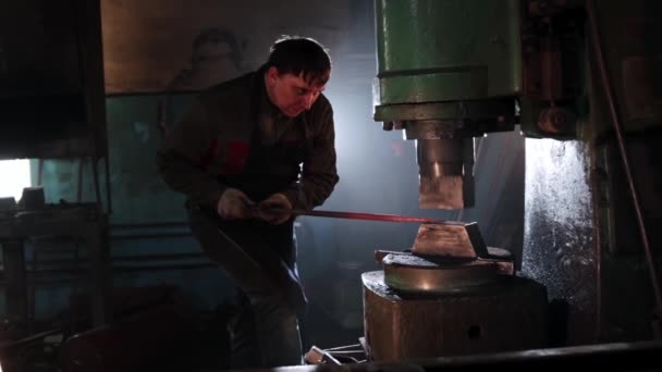 Hombre enfocado herrero adelgazamiento de una pieza de metal oblonga caliente bajo una prensa automática — Vídeos de Stock