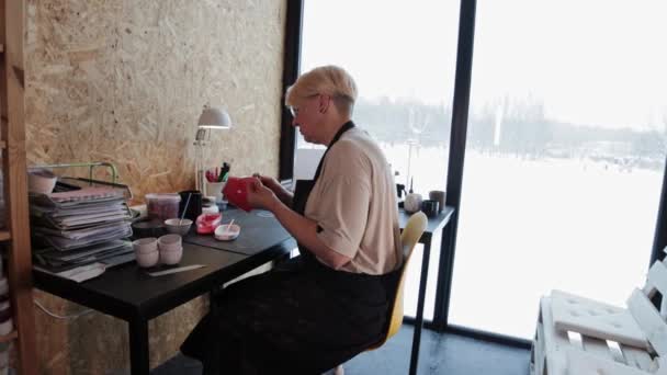 An elderly woman painting ceramic mug with red color — Vídeo de Stock