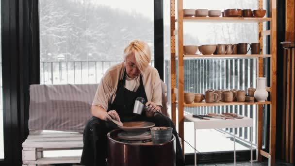 Керамика - пожилая женщина нагревает глиняную тарелку на гончарном круге с помощью фена и удаляет излишки глины с краев — стоковое видео