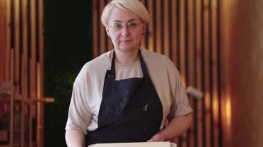 Yaşlı bir kadın ahşap masada kil tabağı gösteriyor.