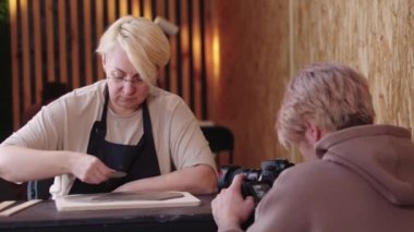 Yaşlı bir kadın ince bir kil parçasını dümdüz ediyor ve kameralı bir adam onu çekiyor.