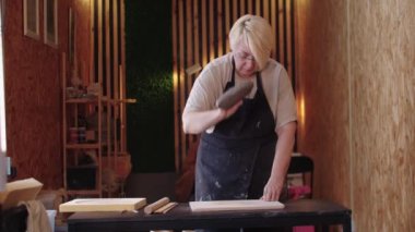 Kısa saçlı yaşlı bir kadın tahtaya bir parça kil atar ve onu düzleştirir.