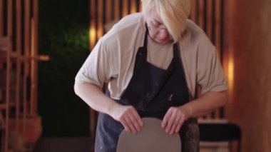 Kısa saçlı yaşlı bir kadın bir parça kili düzleştiriyor tahtaya atıyor ve merdane kullanıyor.