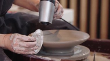 Çömlek tekerindeki kil tabağı saç kurutma makinesiyle yiyen ve düz bir aletle yüzler yapan bir kadın.