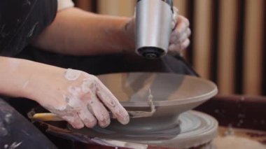 Bir kadın çömlek tekerindeki kil plakayı saç kurutma makinesiyle ısıtıyor ve kenarlarında bir alet kullanıyor.