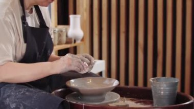 Yaşlı bir kadın çömlek tekerindeki ıslak kil tabakta düz bir alet kullanıyor.