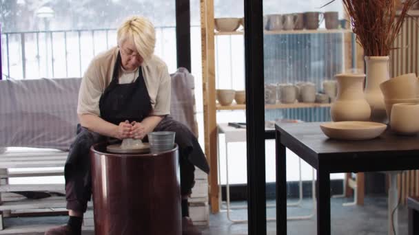 Une femme âgée mouille un morceau d'argile sur une roue de poterie en mouvement dans l'atelier — Video