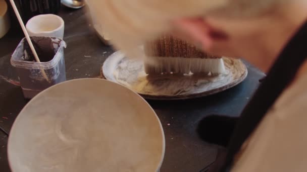 An elderly woman using a big brush to cover new clay plate with special solution — Stockvideo