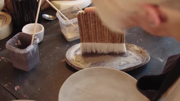 An elderly woman using a big brush to cover new clay plate with brown color paint — Vídeo de stock