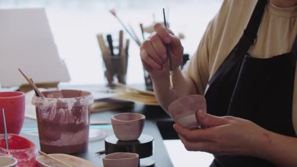Eine ältere Frau bemalt einen kleinen Tonbecher mit staubiger Rosenfarbe — Stockvideo