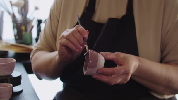 Una donna anziana che dipinge una piccola tazza di argilla con il colore rosa polveroso e guarda nella fotocamera — Video Stock