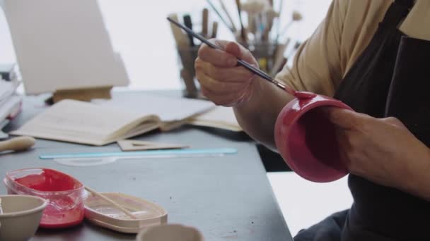 Руки літньої жінки, що малюють глиняний кухоль червоного кольору за допомогою пензлика — стокове відео