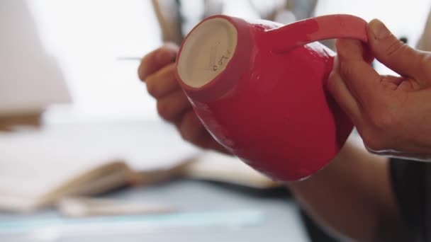 Hands of an elderly woman painting clay mug in red color — Stockvideo