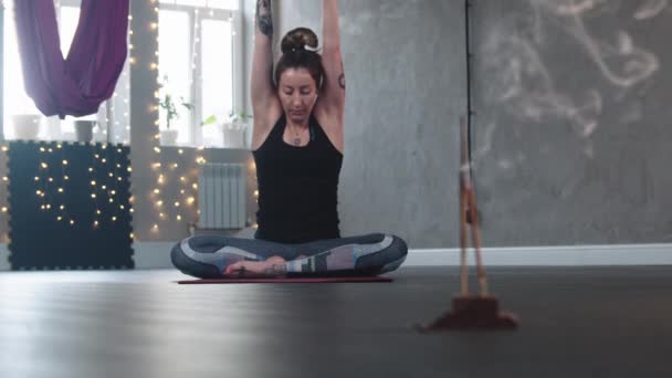 Smoldering incense in the studio - a woman sits on yoga mat and stretching her body — Vídeo de stock