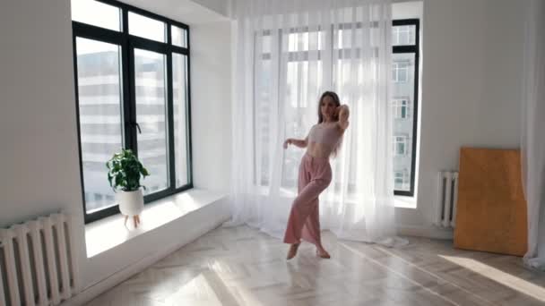 Молода струнка жінка повільно танцює в білій просторій кімнаті біля вікон — стокове відео
