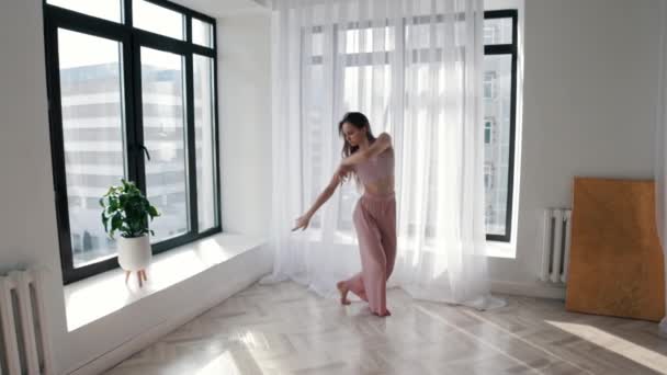 Молода струнка жінка повільно танцює руками в білій просторій кімнаті біля вікон — стокове відео