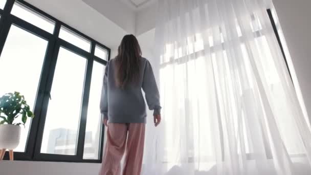 Молода жінка з довгим волоссям йде до вікна і відтягує завісу — стокове відео