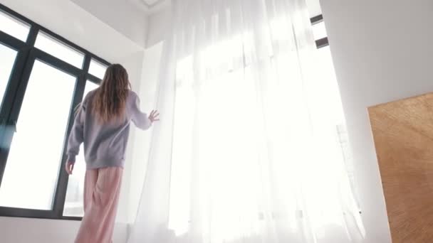 긴 머리를 가진 한 젊은 여성 이 창문으로 걸어 나와 커튼을 뒤로 젖히고 방 안으로 빛을 밝힙니다 — 비디오