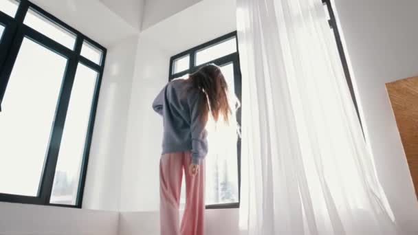 Усміхнена молода жінка танцює перед вікном у яскравому денному світлі — стокове відео