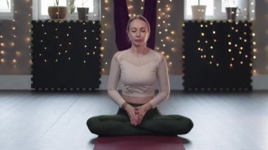 Sarışın bir kadın yoga minderine oturur ve nefes alma egzersizleri yapar.