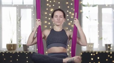 Genç bir kadın yoga hamağında sallanıyor ve avuçlarını birleştiriyor.