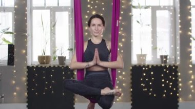 Elleri bir arada yoga hamağında sallanan gülümseyen genç bir kadın.
