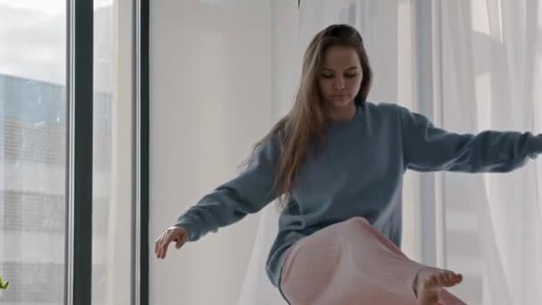 Молодая женщина в пастельной одежде танцует у окна — стоковое видео