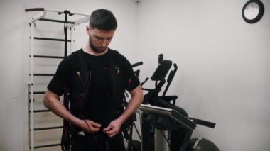 Bir adam acil yardım ekipmanlarıyla bir yelek bağlayıp spor salonundaki kameraya bakar.
