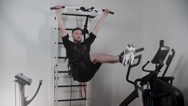 Концентрований чоловік у обладнанні для електростимуляції висить на підставці і тягне ноги вгору — стокове відео