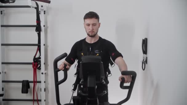 Kjekk, skjeggete mann med elektrostimuleringsdrakt og trening på treningssykkel – stockvideo
