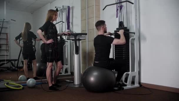 Young man sitting on fitness ball and showing to the woman in ems suit how to do an exercise — Stock Video