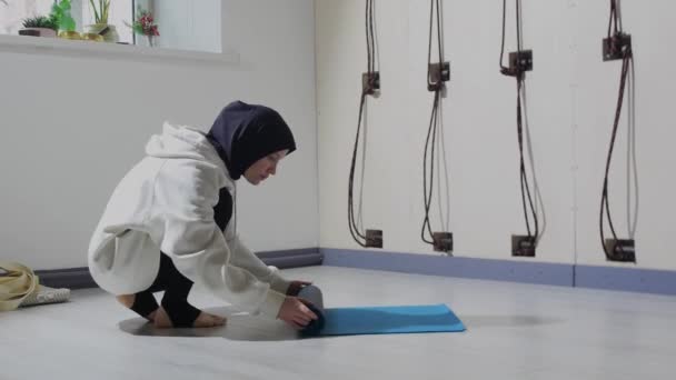 A young woman in hijab picks up the yoga mat from the floor — Stock Video
