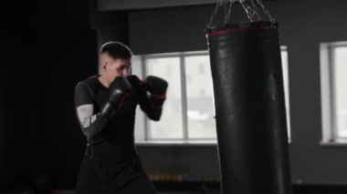 Spor kıyafetleri giyen atletik genç boksör spor salonunda kum torbasına çarpıyor. Orta çekim