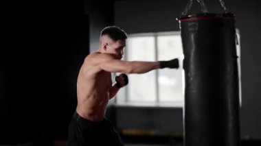 Atletik üstsüz genç boksör kum torbasına vuruyor. Orta çekim