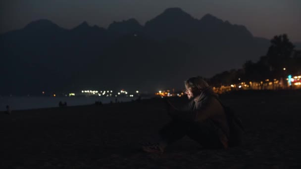 Blondyn Beżowej Bluzie Kapturem Siedzący Nocnej Plaży Sms Ujący Telefonie — Wideo stockowe
