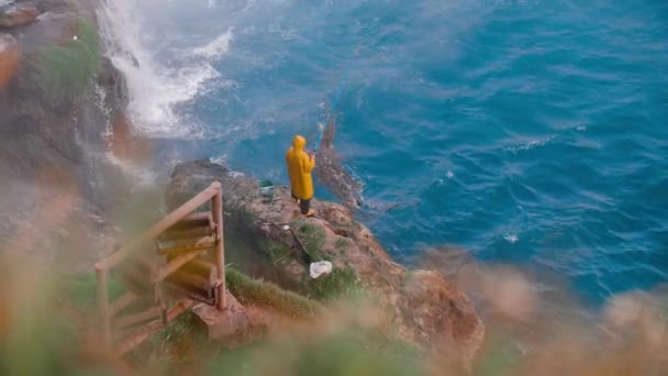Seorang Pria Dengan Mantel Hujan Kuning Sedang Memancing Atas Batu — Stok Video